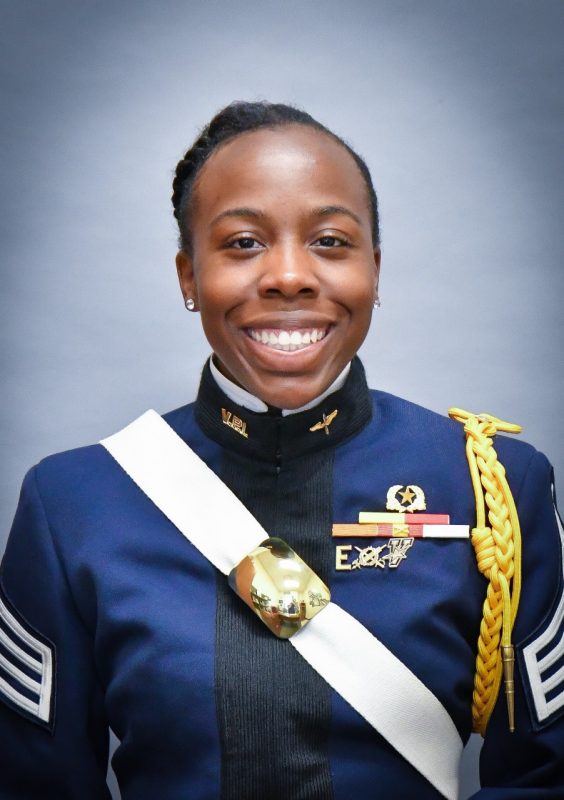 Heron smiles in a portrait shot set against a gray background. She is wearing her dress uniform blouse, white cross belt with shiny brass, and cadet ribbons. She is smiling. 