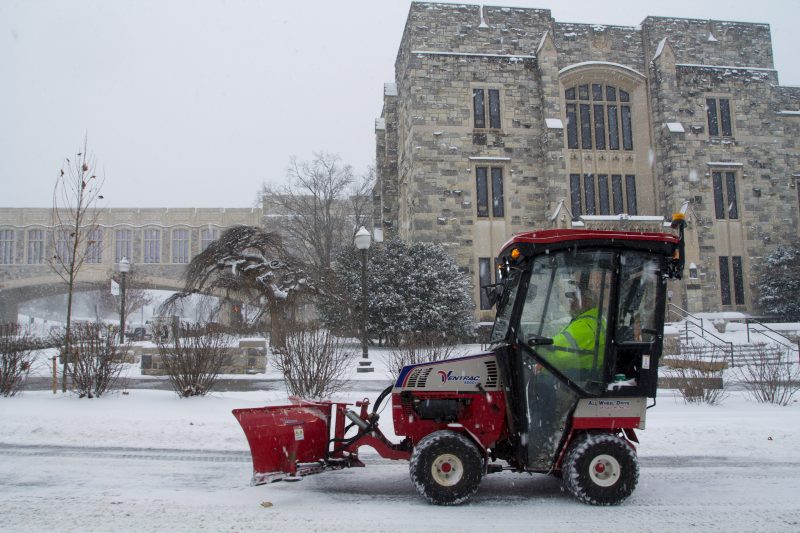 A mini red snow plow removes snow on a roadway in front of grey Hokie Stone Newman Library on a snowy day. 
