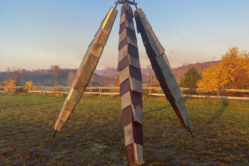 An image of Joe Kelley's sculpture, "Mantis," pictured in a field at the golden hour, fall trees and mountains in the distance.