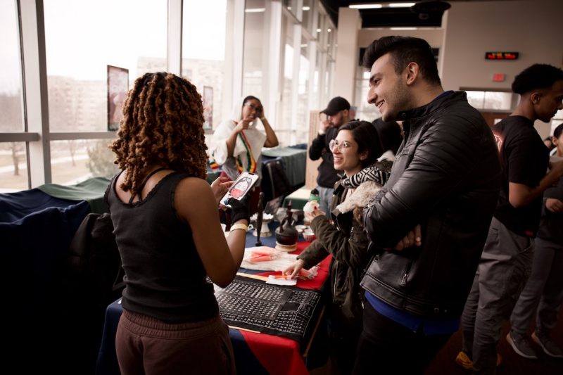 Students dressed for cooler weather smile as they stand in front of a table at International Cafe Hour. The table holds several items of cultural significance, including fabrics and sculptures. Another student with their back to the camera is engaged in conversation with them.. Other students in the background are similarly focused on cultural exhibits.