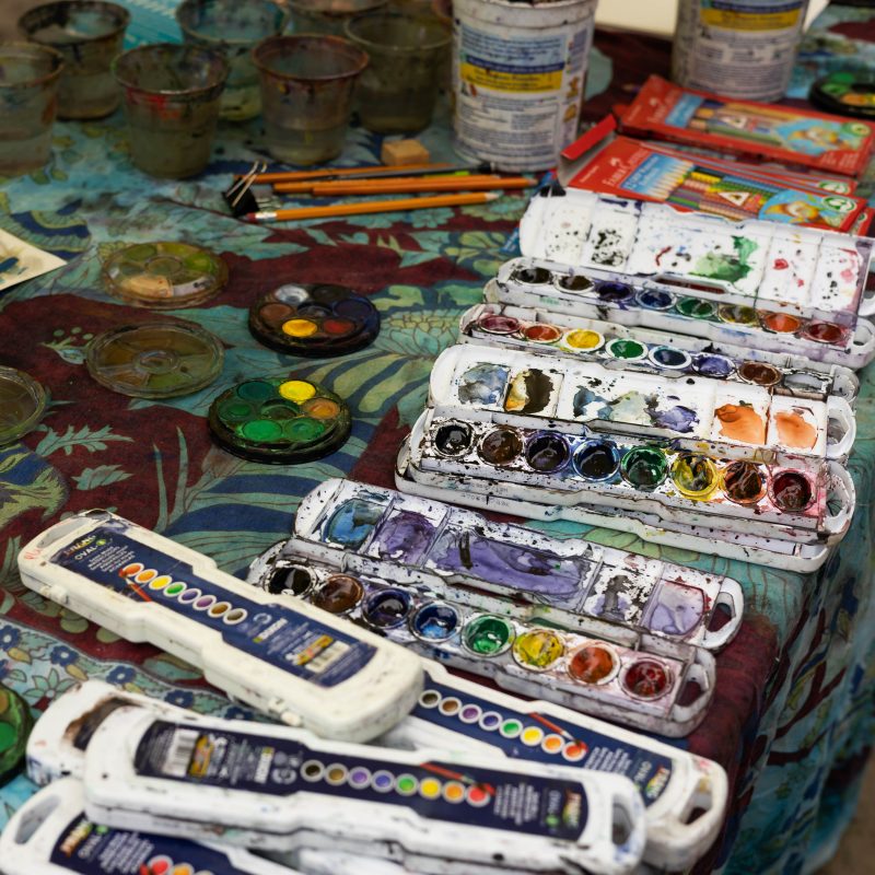 several trays of used multicolor water paint are scattered across a table with a floral tablecloth. In the background are paint-stained cups of water, pencils, and other out-of-focus art supplies.