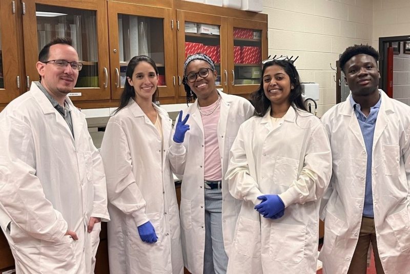 Tim Jarome stands next to four smiling student researchers, all wearing white lab coats, in his Virginia Tech lab.