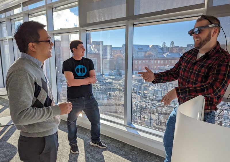 Three people stand in front of a window overlooking Virginia Tech campus. They are (from left) Bo Ji, Brendan David-John, and graduate student Matthew Corbett . The man on the right is wearing an augment-reality device. 