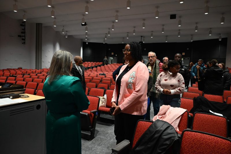 Janice Underwood in a jade satin dress greets a long line of attendees, which extends back into the auditorium. The person at the front of the line is wearing a pink sweater adorned with the letters “A K A” to represent their sorority affiliation.