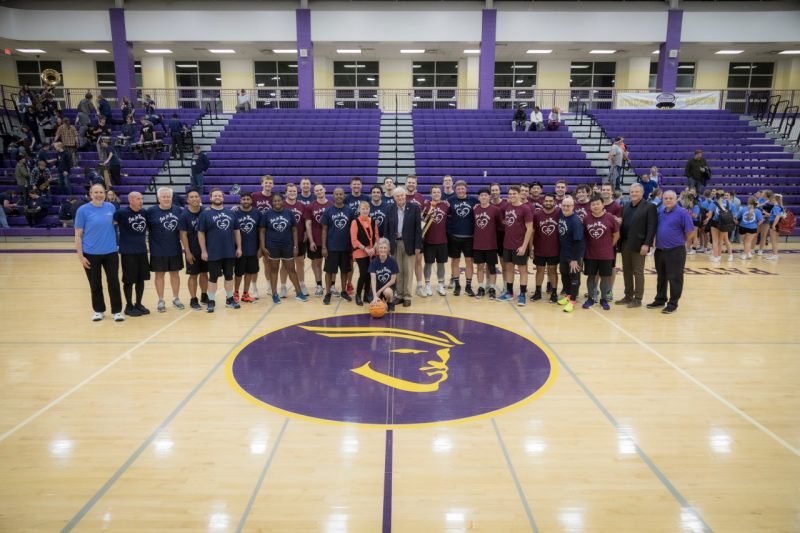 A large group of individuals in blue and maroon t-shirts assemble at midcoast of a basketball court following a game.