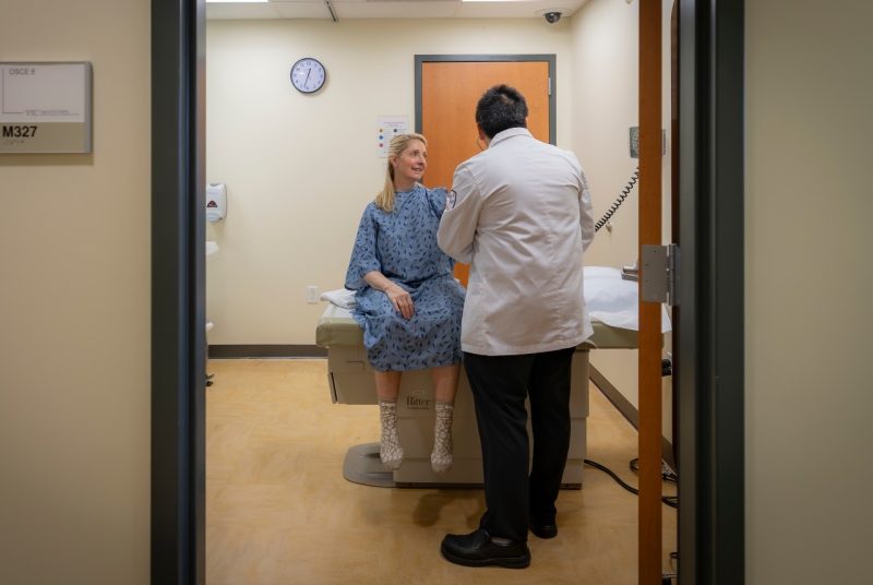Woman wearing an exam gown sits on an exam table. Rear-facing medical student in white coat interacting with her.