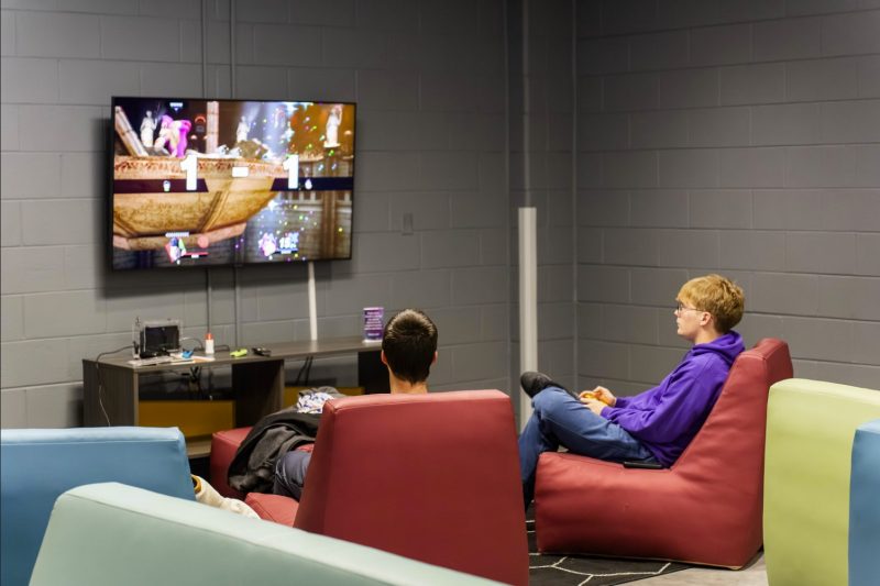 Two students, one in a purple hoodie, sit in red armless chairs in front of a video game station with a wall-mounted TV.