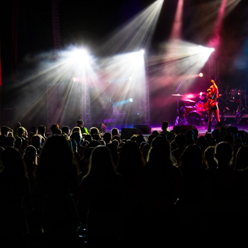 Several bright white lights shine from metal rigging around the stage at Burruss Auditorium, illuminating a crowd of students below with their back to the camera. An out-of-focus artist brightly lit with multicolored lights is performing on stage with an arm raised and a guitar in front of them.