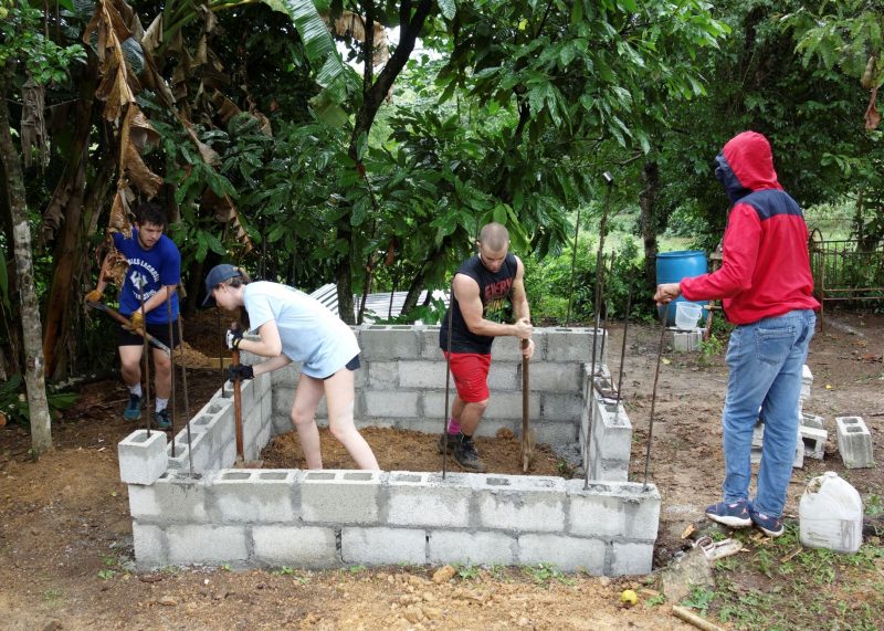 Four students with shovels and other tools work on a structure made of cinderblocks and rebar. They are surrounded by lush greenery with construction materials and containers in the background.