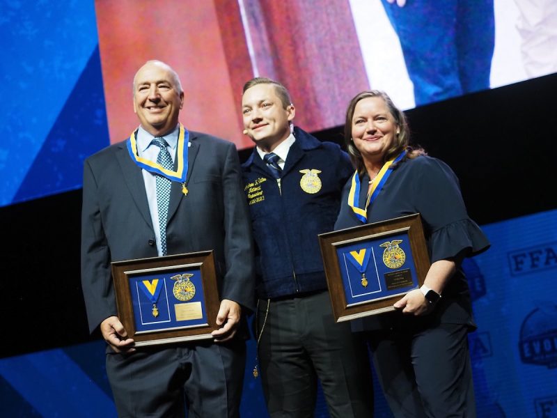 Two men and a woman stand together on stage, holding up award plaques and smiling.