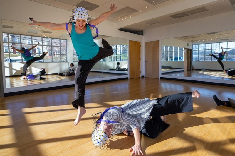 In a dance studio, a woman leaps in the air, while another lies on the floor nearby, kicking her leg out.