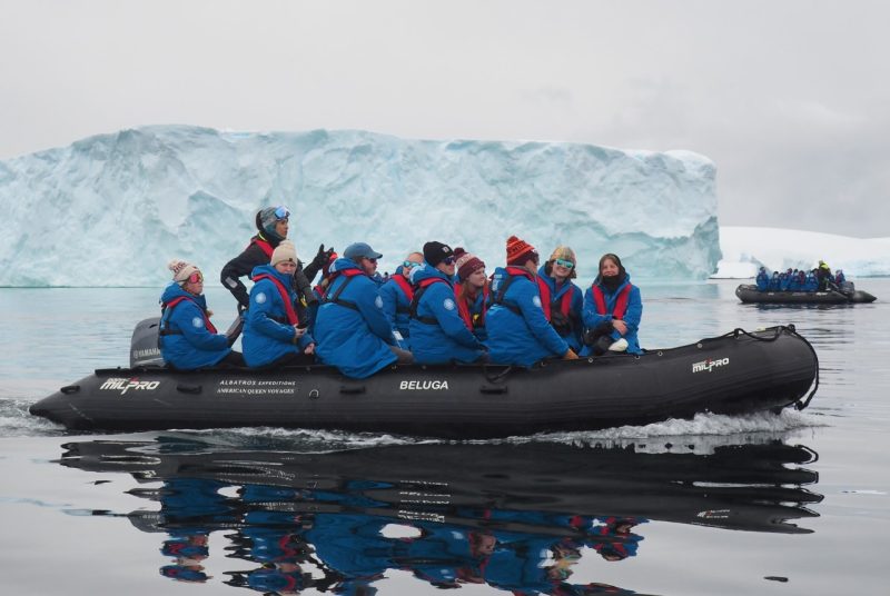 A group of people in an inflatable zodiac boat pass in front of an iceberg.