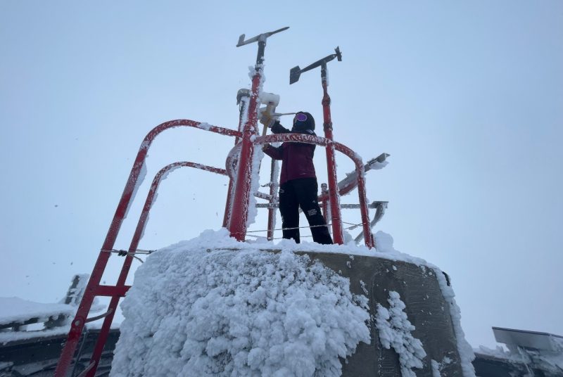 A person in winter clothing uses a tool to remove ice from a weather monitoring instrument.