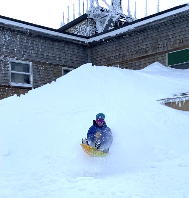 A person sleds down an embankment of snow that has pilled up against a building.