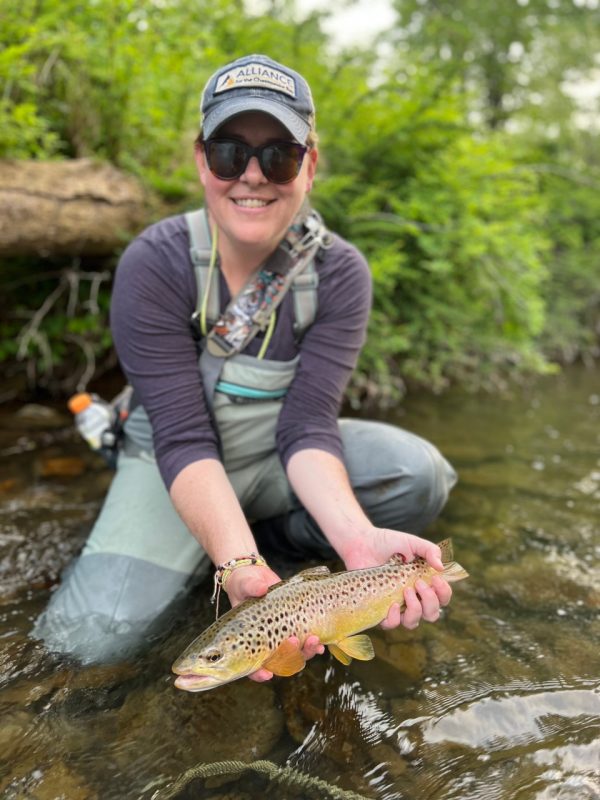 A person kneels in a stream while holding a trout. 