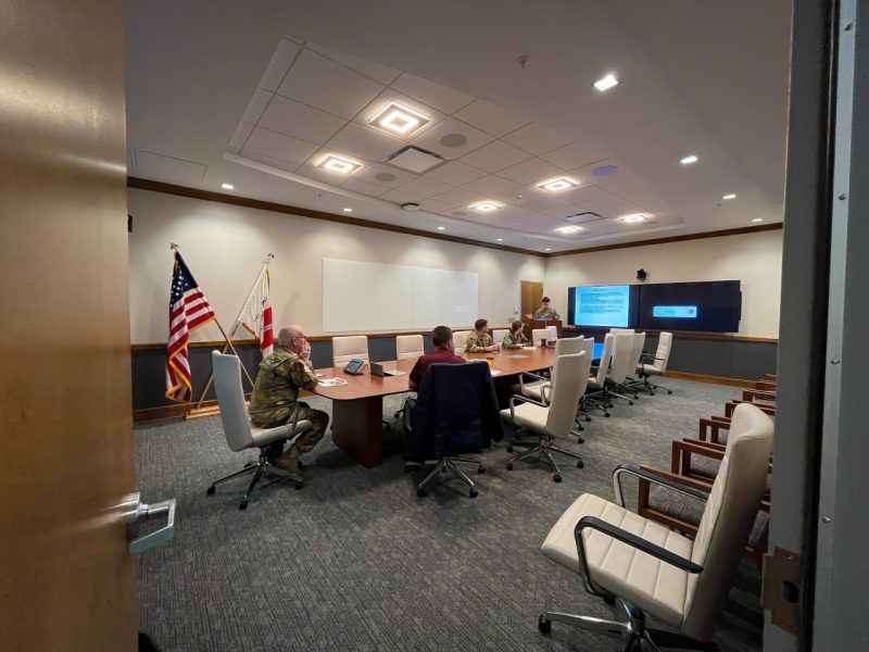  The commandant sits in a large conference room with cadets at the end of the table. One is talking to the commandant from behind the podium as two cadets listen nearby. The team advisor sits next to the commandant.