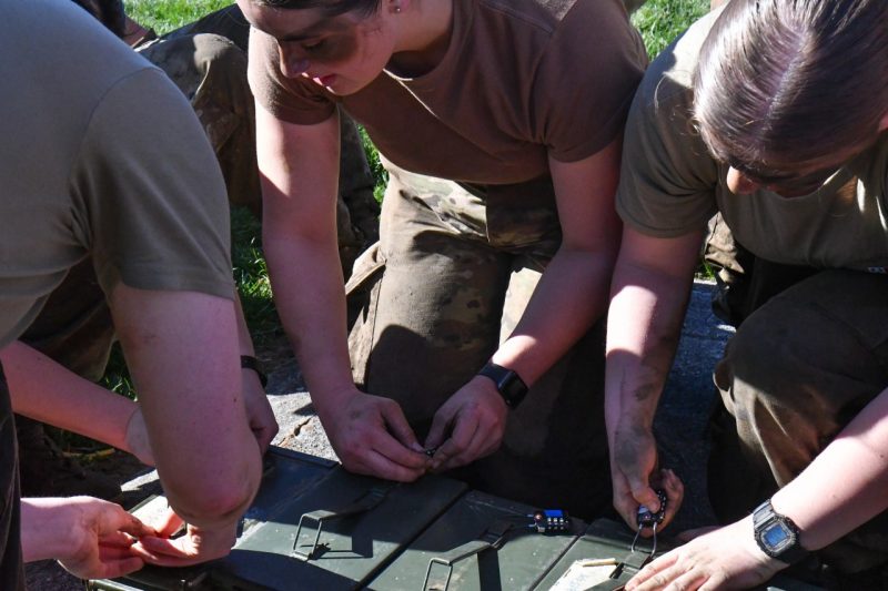 A group of muddy cadets huddle on the ground around locked containers and try to unlock them.