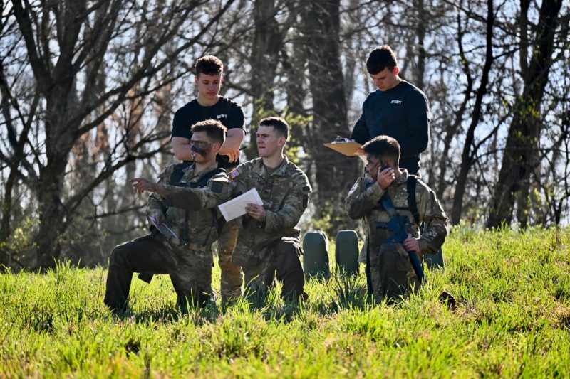  Three cadets in muddy camouflage kneel while two of them discuss a target’s position. The third is listening to the radio while two other cadets grade them on their performance.
