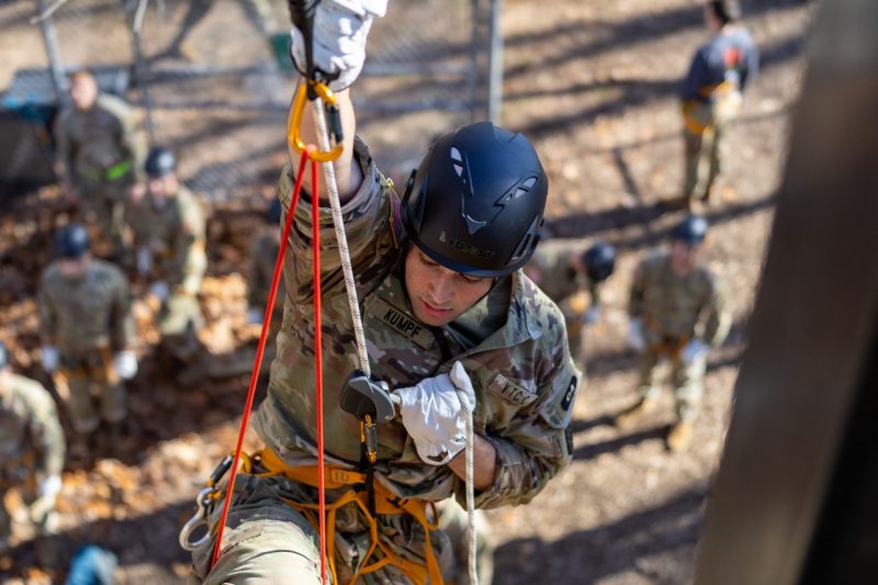  A cadet in climbing gear and a helmet looks down while climbing up the tower. Cadets below him look up as he climbs.