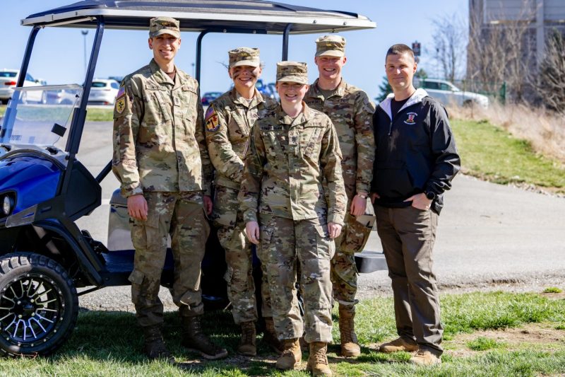 Four cadets in uniform stand with their faculty advisor in front of a golf cart. Everyone is smiling.