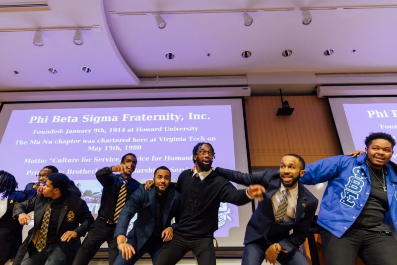 Eight Black fraternity members dance at the front of a Goodwin Hall auditorium