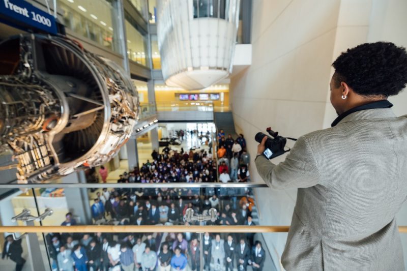 A student aims a camera over a balcony at hundreds of Black men standing in an atrium below.