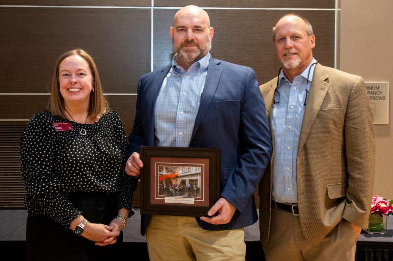 A smiling white woman with two white men, one holding a framed photo award