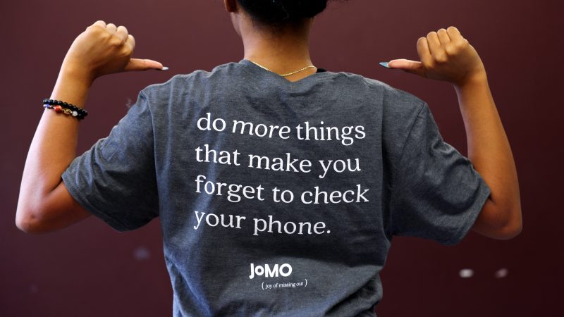 A person facing away points with their thumbs to the back of the tee shirt they are wearing, which reads, "Do more things that make you forget to check your phone. - Joy of Missing Out"