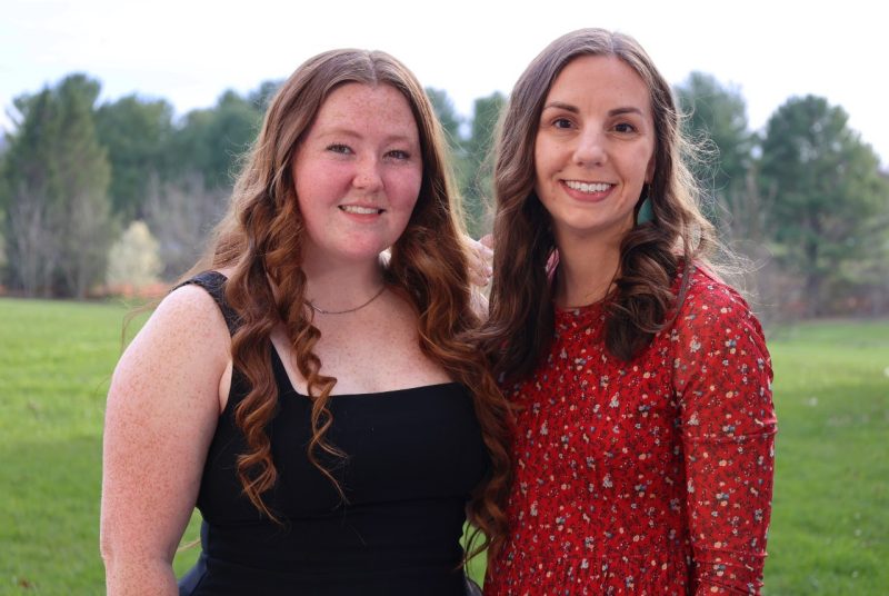 Two people stand in front of a row of trees.  