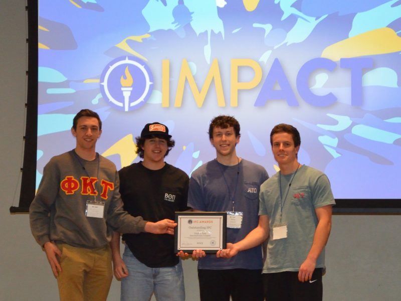 Four students in clothing bearing their different fraternities' letters stand smiling, holding their certificate of award. A screen behind them shows the IMPACT logo.