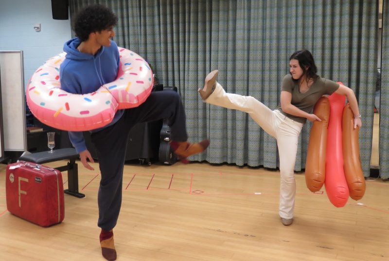 Bianca, played by senior double major in public health and theatre, Aryan Mathur, and Katherine, played by junior double major in theatre and English literature, Virginia Ames Tillar rehearse a fight using inflatable pool floats.  There is a red suitcase on the floor. One float, shaped like a frosted donut, is around Bianca, and the other float is held by Katherine, and is shaped like a hot dog on a bun.