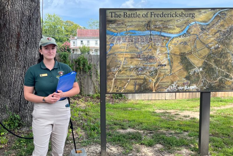A college-aged woman, dressed in a green polo and green hat, stands beside a large outdoor map titled "The Battle of Fredericksburg."