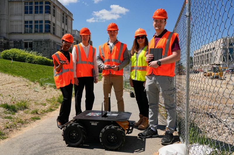 Five people in construction gear smile behind robot and drone.