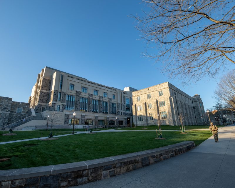 A wide view of the exterior of Holden Hall. The new four story wing is visible and accompanies  a three story wing.  A cadet walks by on the path outside the building.