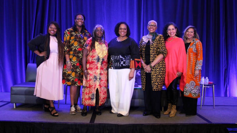 Leadership Panel FWCA 2024 A group of women pose against a blue backdrop wearing bright colors and smiling. Behind them are chairs set up for a panel discussion on a stage.
