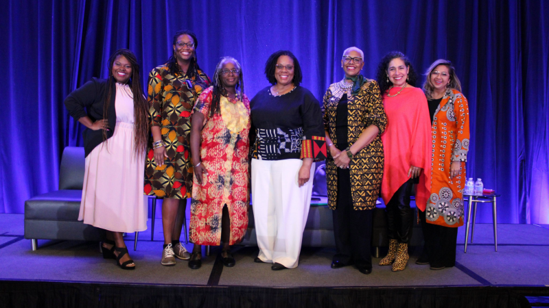 A group of women pose against a blue backdrop wearing bright colors and smiling. Behind them are chairs set up for a panel discussion on a stage.