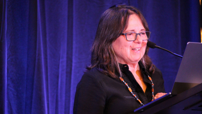 Cherrie Moraga at FWCA 2024 A Latine woman with graying shoulder-length hair and glasses stands against a blue backdrop and presents from a podium and laptop. She is wearing a colorful beaded necklace.