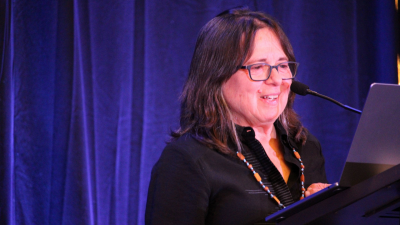 A Latine woman with graying shoulder-length hair and glasses stands against a blue backdrop and presents from a podium and laptop. She is wearing a colorful beaded necklace.