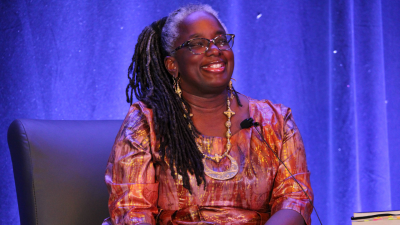 A Black woman with gray hair at her roots and dark locks smiles while seated giving a presentation. She is wearing a bright orange and gold dress and hand-made jewelry.