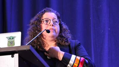 Gabrielle Tayac at FWCA 2024 An Indigenous woman with long curly hair and glasses holds one hand to her throat while she presents from a podium and laptop. Her jacket has three stripes in pink, orange, and white.