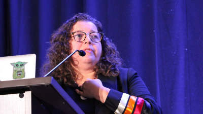 An Indigenous woman with long curly hair and glasses holds one hand to her throat while she presents from a podium and laptop. Her jacket has three stripes in pink, orange, and white.