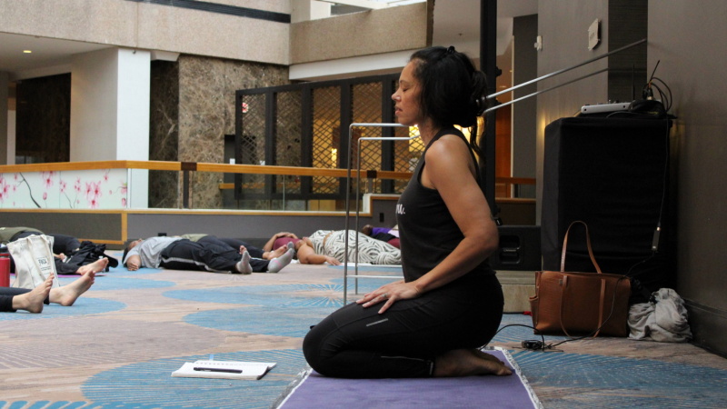 A woman in black yoga gear kneels on her yoga mat with her eyes closed. Many women take rest poses on their mats around the room in the background.