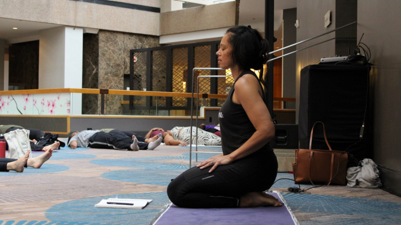 Lacretia "Cre" Dye teaches yoga at FWCA 2024 A woman in black yoga gear kneels on her yoga mat with her eyes closed. Many women take rest poses on their mats around the room in the background.