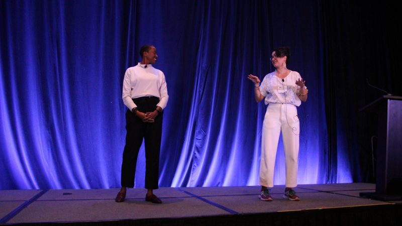 Angela's Pulse at FWCA 2024 Two people in stylish clothing stand a few feet apart looking at each other as they explain something to the audience beyond the stage. Both are smiling.