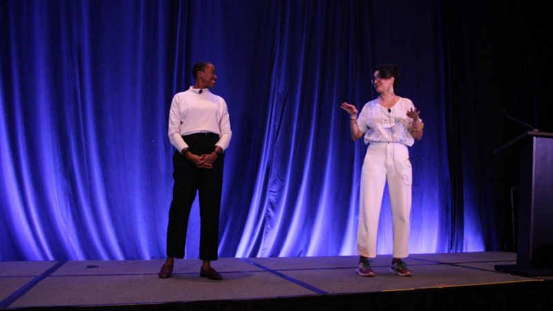 Two people in stylish clothing stand a few feet apart looking at each other as they explain something to the audience beyond the stage. Both are smiling.