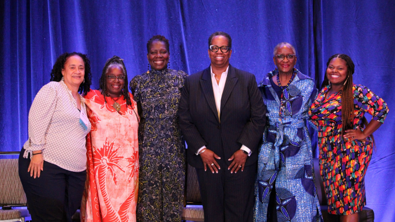 A group of women pose against a blue backdrop. The woman in the center, who is the primary subject, is wearing a suit and glasses.