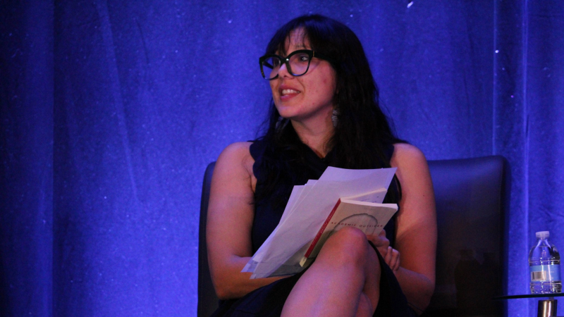 Victoria Reyes at FWCA 2024 A Latine woman with long dark hair and glasses sits in a leather chair and looks out into the audience behind the camera. She holds a book and several pages in her hands.