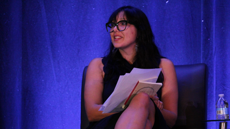 A Latine woman with long dark hair and glasses sits in a leather chair and looks out into the audience behind the camera. She holds a book and several pages in her hands.