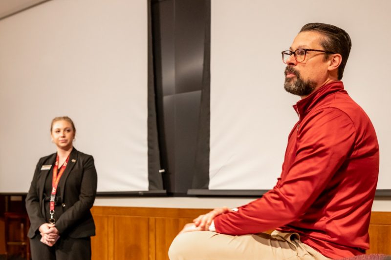 A man in a red shirt sits on a table and talks while a young woman in a black suit stands in the background listening.