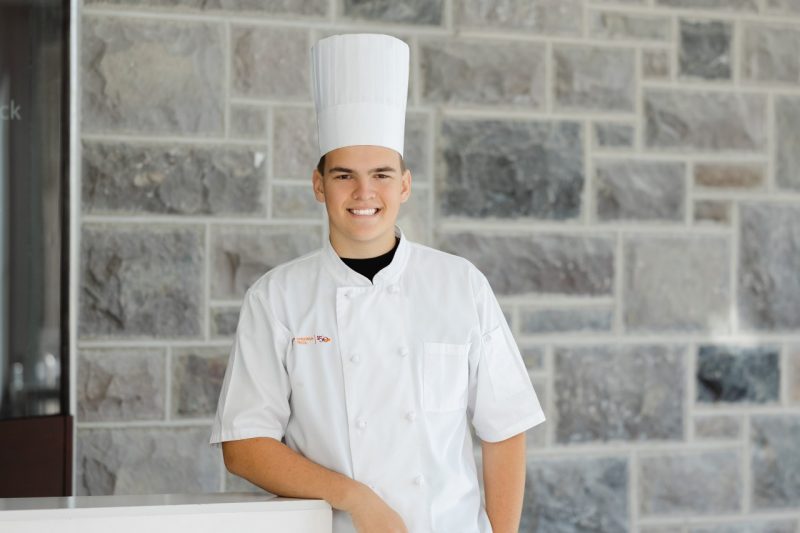 Headshot of Will Racek wearing a chef's hat and uniform and standing in front of a Hokie Stone wall.
